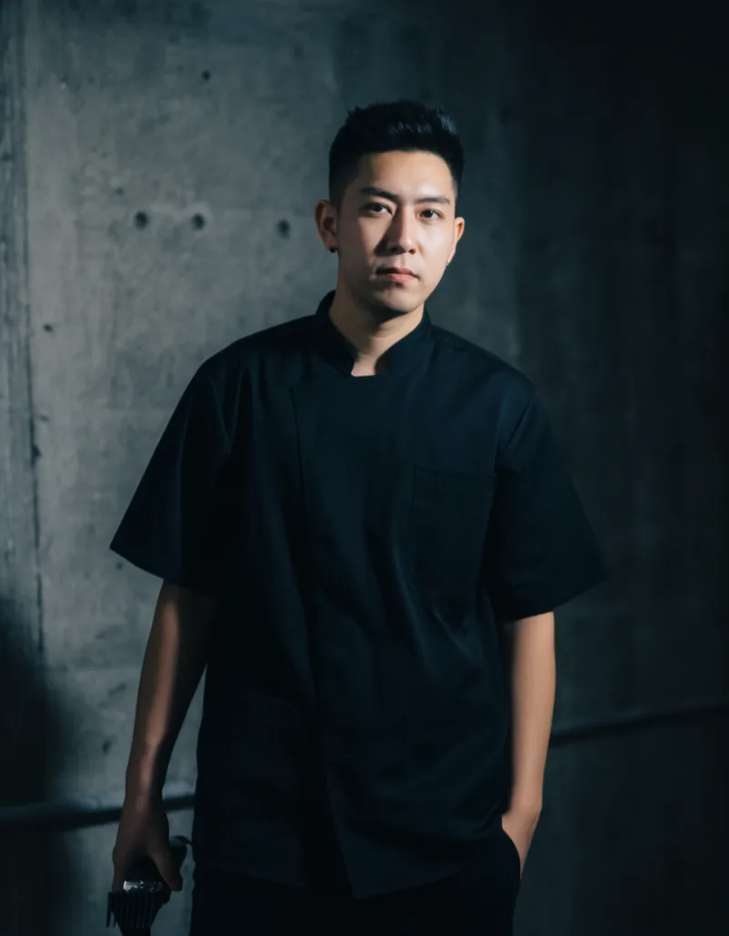 Portrait of Milan Petrov, a barber in his early thirties with close-cropped dark hair in a plain black work shirt, standing in the concrete studio