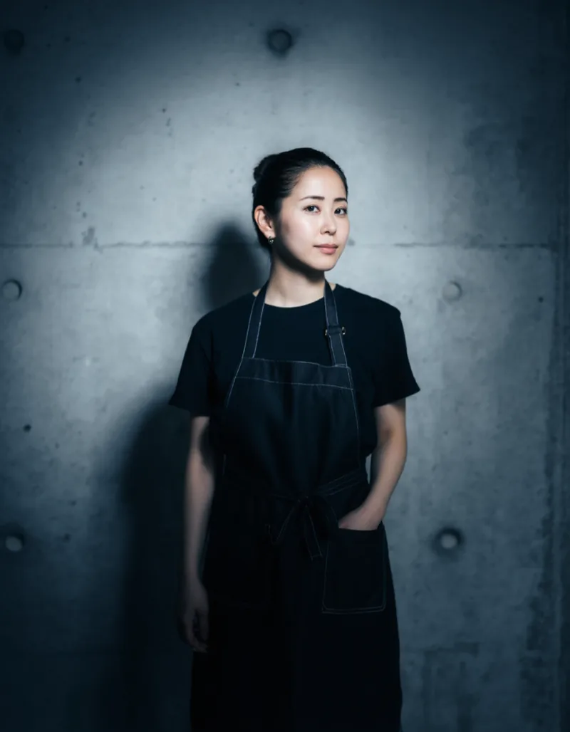 Portrait of Jule Radek, a barber in her late twenties with her hair tied back, wearing a charcoal work apron in the concrete studio