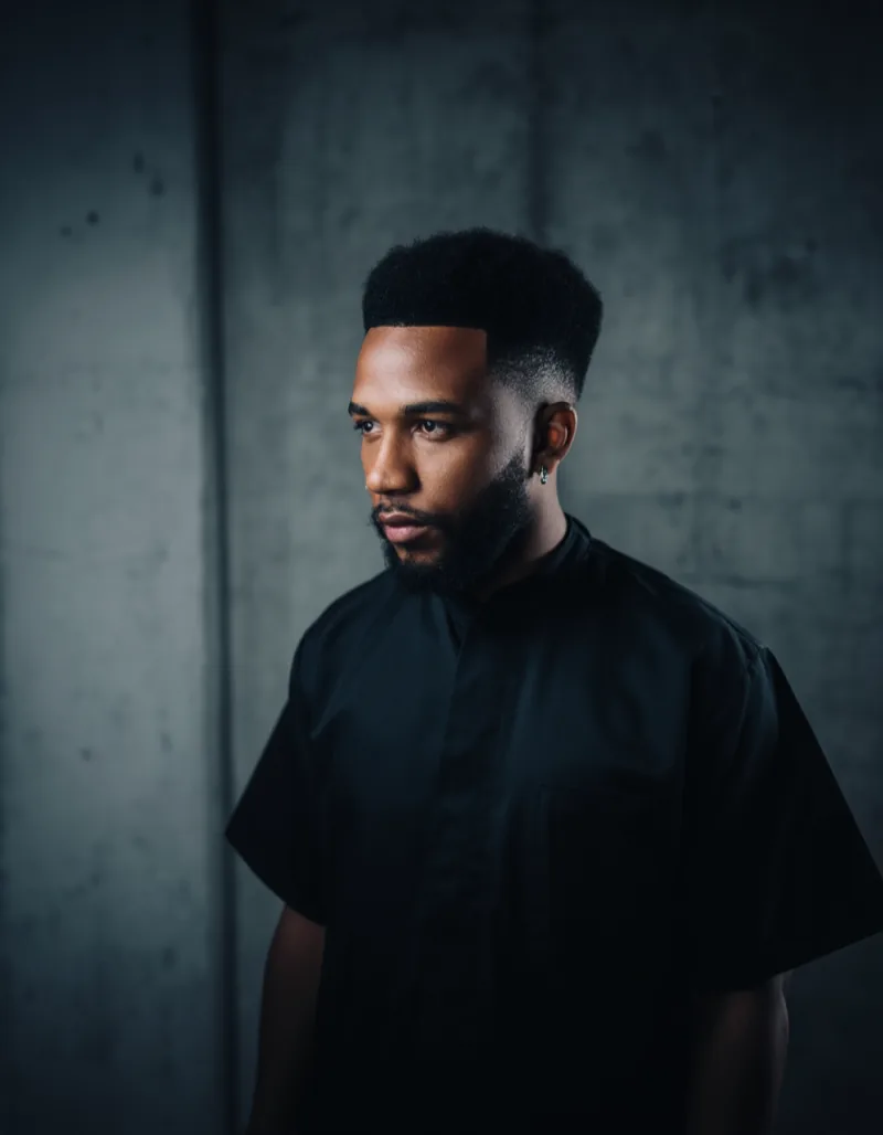 Portrait of Kwame Osei, a barber in his thirties with a short afro fade and trimmed beard, wearing a black work shirt in the concrete studio